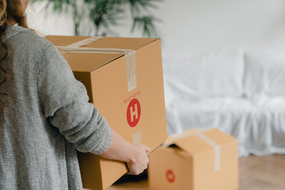 A person wearing a grey knitted sweater is holding a large cardboard box sealed with clear packing tape during a home relocation process inside a well-lit room. The box features a red 'H' logo and is being prepared for transportation, with their left hand supporting the bottom while their right hand holds a box cutter or tape dispenser. In the background, there are additional cardboard moving boxes, some open, placed on the floor, along with a white, cushioned sofa against a wall, indicating a typical living space being prepared for furniture transport. The environment includes a wooden floor and natural light illuminating the scene, aligning with professional removals and packing activities undertaken by Man With a Van Hendon as part of a house removals or moving service in Hendon.