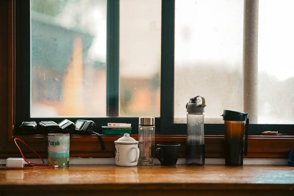 A daytime view of a wooden windowsill inside a house, featuring various household items including a small stack of black remote controls, a green and white cardboard cup, a white ceramic mug, a clear glass water bottle, a black ceramic mug, a large plastic water jug with a handle, and a brown travel mug with a lid. Behind the window, an out-of-focus outdoor scene is visible with blurred shapes and muted colors. The Natural light coming through the window highlights the arrangement of these items, which are positioned for packing or temporary storage during a home relocation process. This interior setting, with its focus on household objects, aligns with furniture transport and packing aspects of house removals, supported by the presence of a company such as Man With a Van Hendon specializing in removals and moving logistics.