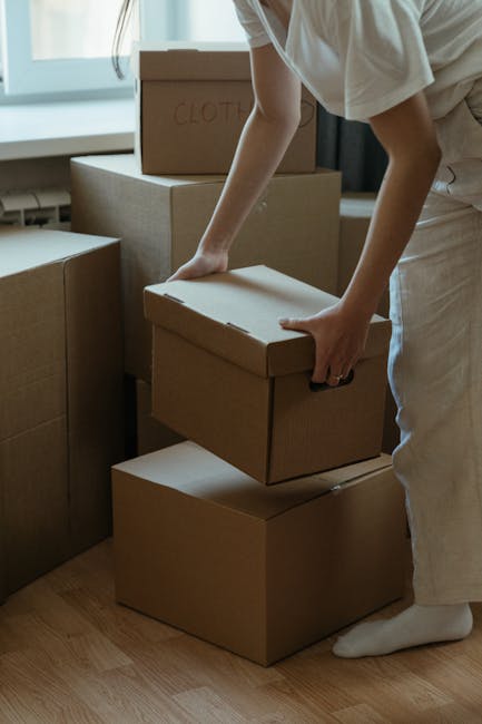 A person wearing a grey knitted sweater is holding a large cardboard box sealed with clear packing tape during a home relocation process inside a well-lit room. The box features a red 'H' logo and is being prepared for transportation, with their left hand supporting the bottom while their right hand holds a box cutter or tape dispenser. In the background, there are additional cardboard moving boxes, some open, placed on the floor, along with a white, cushioned sofa against a wall, indicating a typical living space being prepared for furniture transport. The environment includes a wooden floor and natural light illuminating the scene, aligning with professional removals and packing activities undertaken by Man With a Van Hendon as part of a house removals or moving service in Hendon.