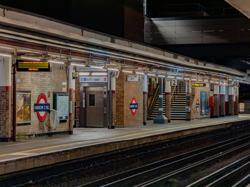 Inside Harrow and Wealdstone Tube Station platform, illuminated by artificial lighting, the scene shows brick walls adorned with London Underground signage, including a 'Harrow & Wealdstone' roundel a
