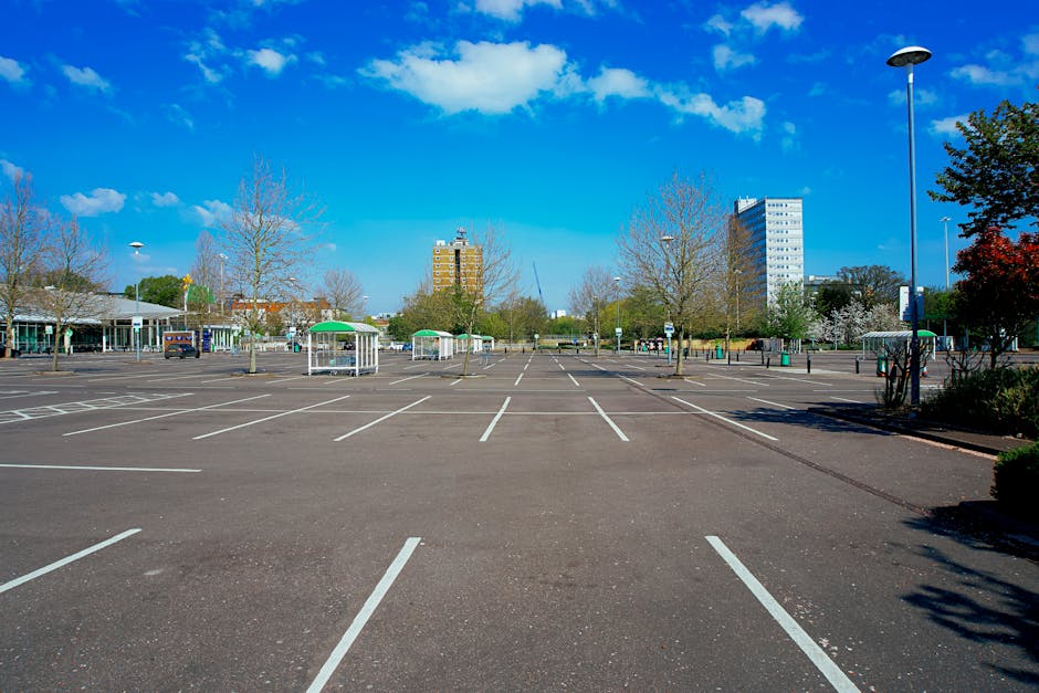 A large, mostly empty parking lot with clearly marked white parking spaces on smooth asphalt, surrounded by leafless trees and a few small shelters with green roofs. In the distance, tall office or apartment buildings are visible against a bright blue sky with scattered clouds. A few cars are parked near the shelters, and there are street lamps along the edges of the parking area. The scene suggests a quiet, open space appropriate for vehicle loading and unloading, such as during a house removal or furniture transport operation. Man With a Van Hendon, a removals service, may use this parking lot for loading or staging during home moves, with the area offering ample space for loading vans and managing packing materials while facilitating efficient relocation logistics.