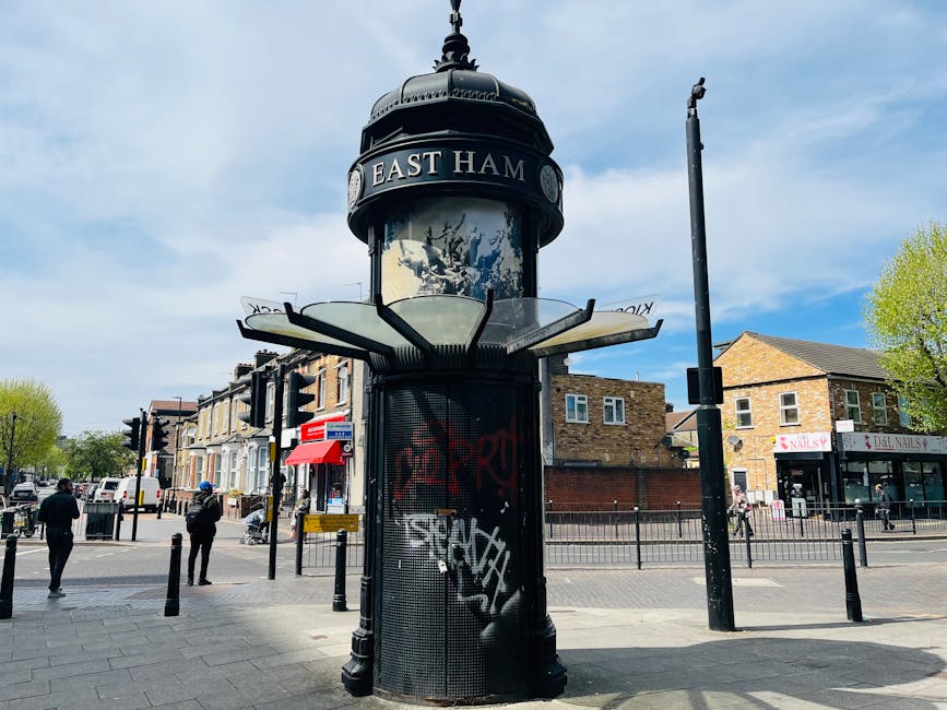 A historic cylindrical street clock with a black metal casing and a domed top, displaying the words 'East Ham' on a sign near the roof, is situated on a busy street corner. The clock face is partially obscured behind a glass shield with decorative metalwork, and below it, a black metal grille covers an interior section possibly housing electronic components. Graffiti tags are visible on the lower part of the clock casing. Surrounding the clock, there are pedestrians walking on the sidewalk, some wearing jackets, and various storefronts including a nail salon and a hairdresser are visible in the background. To the right, there is a tall, empty streetlight pole, and on the left, a pedestrian crossing with black metal bollards separating the sidewalk from the road. The scene is set on a partly cloudy day with sunlight illuminating the street scene, including trees with fresh green leaves, indicating spring or early summer. The environment and the urban setting suggest this may be part of a typical home relocation or moving logistics scene often seen in busy town centers, supported by local amenities and infrastructure, such as the nearby shop fronts and pedestrian activity. Man With a Van Hendon’s removals services are implied through the contextual setting.