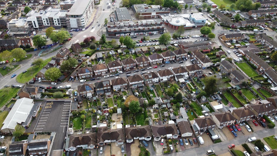 An aerial view of a residential neighbourhood showing rows of terraced houses with small backyards, some featuring sheds and garden furniture. Several houses have driveways with parked cars, and narrow pathways lead to rear gardens. Surrounding the houses, there are patches of green lawns, trees, and narrow alleyways. In the top left corner, a large parking area with marked bays is visible, adjacent to a modern commercial building with flat roofs and multiple windows. The scene is captured during daylight with clear weather, and the overall image illustrates a typical London suburb environment. This context relates to house removals and home relocation services offered by Man With a Van Hendon, involving furniture transport and packing during moves from property to property.