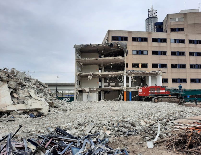 A partially demolished multi-storey building with three floors, exposing interior walls, support beams, and scattered debris on the site. In the foreground, there is a large pile of rubble consisting of concrete fragments, broken bricks, and twisted metal. To the right, a red tracked excavator is positioned on the debris, likely used for tearing down the structure during the demolition process. The background features a modern, multi-paneled office building with numerous rectangular windows, and an overcast sky overhead. The scene illustrates a construction and demolition site, where building materials are being carefully dismantled and removed in preparation for new development or relocation activities, aligning with professional house removal and transportation services provided by companies like Man With a Van Hendon.