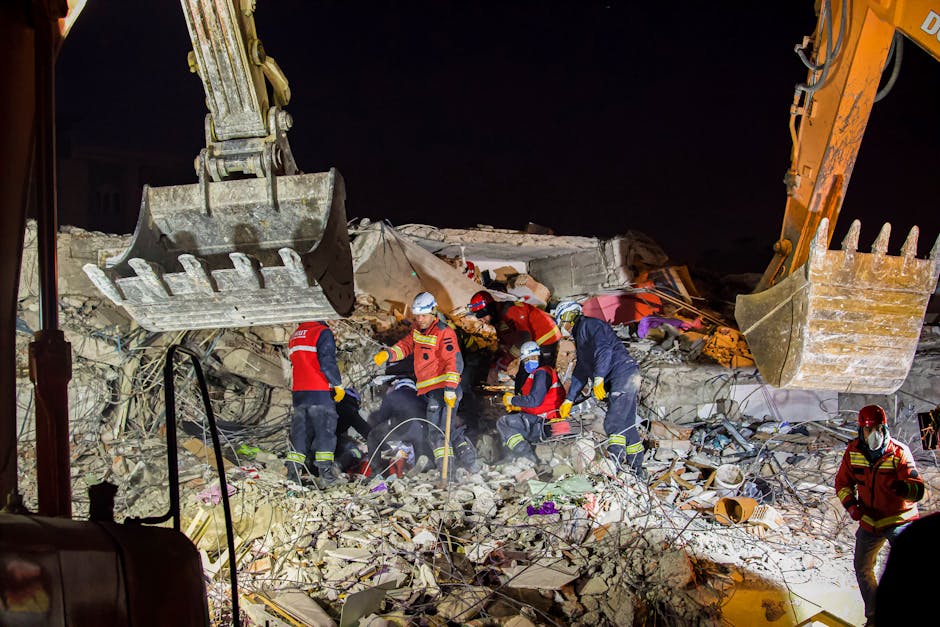 Nighttime scene showing rescue workers in high-visibility jackets and helmets amidst rubble from a collapsed building, with broken concrete, twisted metal, and debris scattered across the ground. Several workers are engaged in clearing debris using tools, while others stand nearby, illuminated by artificial lighting. Large excavator buckets are visible overhead, positioned above the workers, indicating ongoing heavy lifting as part of the emergency rescue and clearance operation. The environment suggests urgent activity related to structural collapse, with dust and broken materials filling the scene, emphasizing the complex and hazardous nature of the work. The setting appears to be an active site for rapid response in a major incident, consistent with emergency clearance services in Hendon, as provided by companies like Man With a Van Hendon, specializing in urgent removals and disaster site clearance.