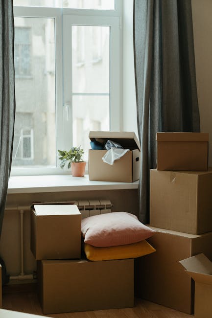 A collection of cardboard boxes of various sizes positioned on a wooden floor near a large, white-framed window, with some boxes stacked and others placed on the floor. One box is open with white packing paper or tissue protruding, indicating packing or unpacking activity related to home relocation or moving. Additional items include a pink cushion and a yellow cushion next to the boxes, with a small potted plant on the window sill behind them. The window allows natural light into the room, which has grey curtains hanging alongside. Visible behind the window are blurred exterior building details. The scene suggests a house moving process, with boxes prepared for transport by a professional removals company, such as Man With a Van Hendon, supporting space-efficient furniture transport and packing and moving services.
