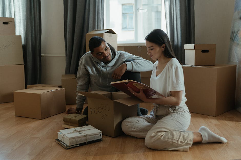 Inside a room with wooden flooring and natural light from a window, a man and a woman are engaged in a home relocation process. The woman is sitting on the floor, dressed in light-colored casual clothing, holding an open book, and surrounded by various cardboard boxes labeled with handwritten indicators such as 'BOOKS.' The man, wearing a gray hoodie, is leaning on his elbow, closely observing the box contents. Several packed boxes of different sizes are arranged around them, some sealed with tape and others partially open. A small stack of wrapped books tied with string is placed near the boxes. In the background, there are additional cardboard boxes and a window with dark curtains, suggesting an ongoing packing or furniture transport activity. This scene reflects the packing phase of a house move coordinated by a professional removals service, with a focus on careful handling of belongings before loading onto a vehicle for transport.