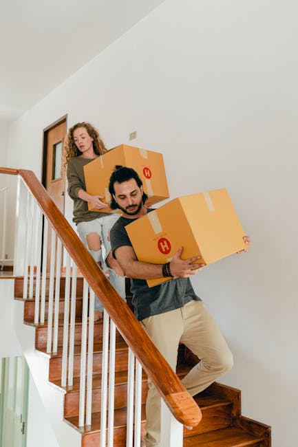 A person inside a well-lit room with large windows, using a box cutter to open or seal a medium-sized cardboard box during a home relocation process. Several similar closed and opened cardboard boxes