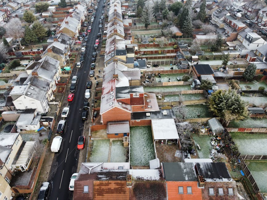 An aerial view of a residential street during winter, showing rows of terraced houses with pitched roofs, some of which have a dusting of snow. The street is lined with parked cars along both sides, including a white van and various small vehicles. Gardens behind the houses are enclosed by wooden fences, with some featuring outdoor furniture, sheds, and decorative plants. The surroundings include trees with sparse foliage, and the overall scene suggests a quiet neighbourhood. This image captures the typical environment involved in home relocation and furniture transport processes, relevant to the services offered by Man With a Van Hendon within their house removals and moving logistics offerings.