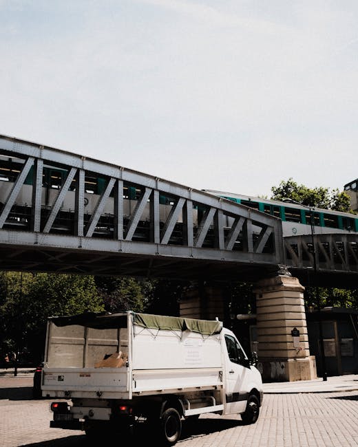 A white moving van with a partially open tailgate is parked on a paved area beneath an elevated railway track supported by stone pillars. The van's cargo area is covered with a green tarp, and some cardboard boxes are visible inside. Against the backdrop of the railway bridge, which is constructed from metal with diagonal bracing, a green train is passing overhead. The surrounding environment includes trees and a building adjacent to the loading area. This scene depicts a home relocation process involving furniture and box transportation, typical of professional removals services such as those offered by Man With a Van Hendon, during the loading and transport phase of a house moving project.