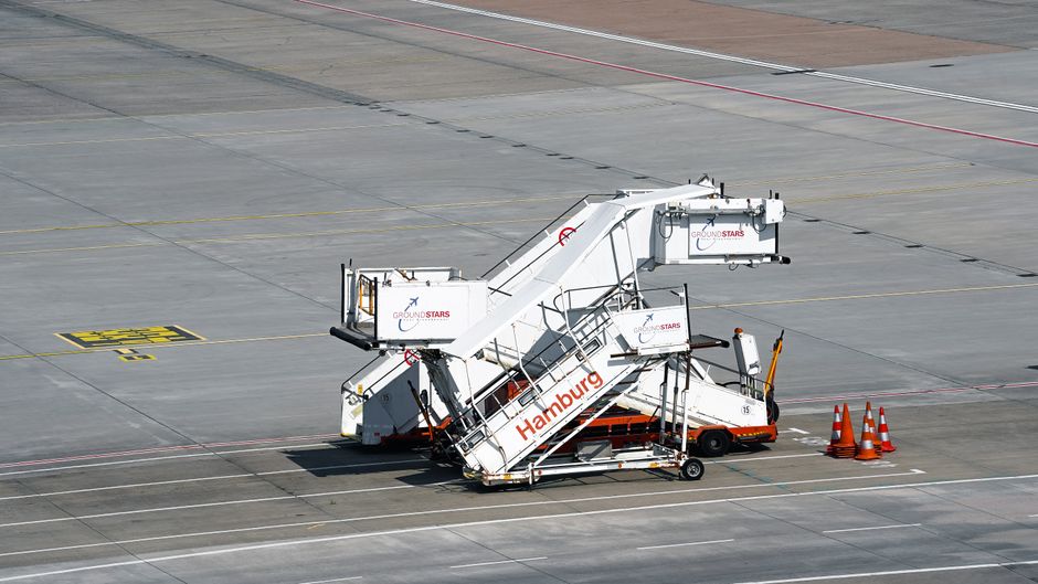 An airport ground handling vehicle with a white and orange exterior is parked on a tarmac, equipped with an extendable passenger stairs marked with the word 'Hamburg' and signage for 'Ground Stars.' The stairs are in a partially elevated position, with black handrails along the sides, and are surrounded by several orange safety cones placed on the ground nearby. The tarmac features yellow and white markings indicating parking positions and guiding lines, with a flat, expansive surface extending into the background. The vehicle is used for aircraft passenger boarding and deplaning, representing an essential part of ground transport logistics at the airport, and is situated in an outdoor area with natural lighting during daylight hours. This scene relates to professional airport ground services, which are vital during aircraft loading and unloading operations, integral to the overall movement process involved in home relocation and furniture transport logistics, as coordinated by companies like Man With a Van Hendon.