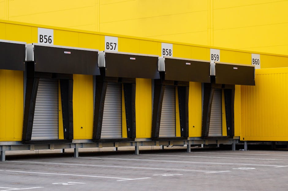 A row of five large, black and grey container shutters, each labeled with a white sign displaying B56, B57, B58, B59, and B60, are mounted on a bright yellow exterior wall of a building. The shutters are partially closed, indicating a loading bay or storage area, with metal supports visible underneath. The ground is paved with asphalt, with white parking markings in the foreground. The bright lighting suggests natural daylight. This scene illustrates a commercial or warehouse environment suitable for home relocation loading and furniture transport services, with the exterior space prepared for moving activities. Man With a Van Hendon offers professional removals, including packing and loading processes, and this loading bay setup supports efficient home relocation operations.