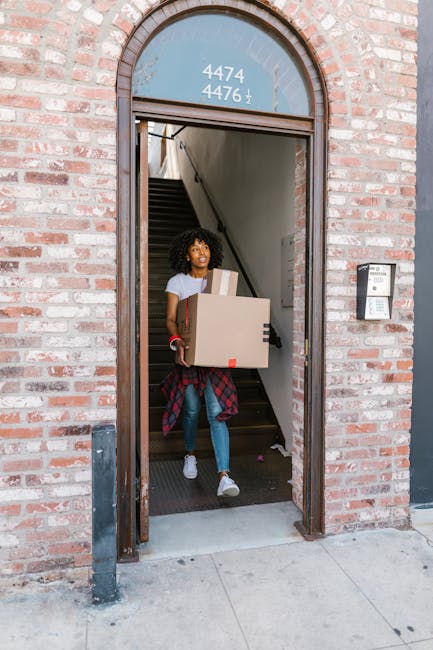 A young woman stands at the entrance of a brick building, holding a cardboard box and a smaller package during a home relocation process. She is positioned inside the doorway, which features an arched window above the doorframe displaying the numbers '4474 4476'. The woman is dressed in casual clothing, including a white t-shirt, jeans, and white sneakers, with a red plaid shirt tied around her waist. Behind her, a narrow staircase with dark carpeting leads upward inside the property, partially visible through the open door. The exterior surroundings include a concrete pavement, a metallic bollard on the left, and a modern intercom panel mounted on the brick wall to the right of the door. The scene captures the logistics of furniture transport and packing during a removals service, with natural lighting highlighting the activity and interior features.