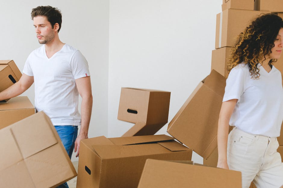 A man and a woman are involved in a home relocation process indoors, surrounded by stacked cardboard boxes of various sizes. The man, wearing a white t-shirt and blue jeans, is lifting or moving a medium-sized cardboard box, with his arm extended and a focused expression. The woman, dressed in a white top and beige trousers, stands nearby with her arms relaxed, looking down at the boxes or the floor. Behind them, there are more cardboard boxes neatly stacked against a plain white wall, some sealed with packing tape. The scene appears to take place in a spacious room, possibly a living area, with ample natural lighting illuminating the space, supporting a professional moving or packing operation conducted by Man With a Van Hendon. The image captures the logistics involved in furniture transport and packing during a professional house removals or estate essentials service, highlighting the careful handling and organization essential for a smooth move into a new home.