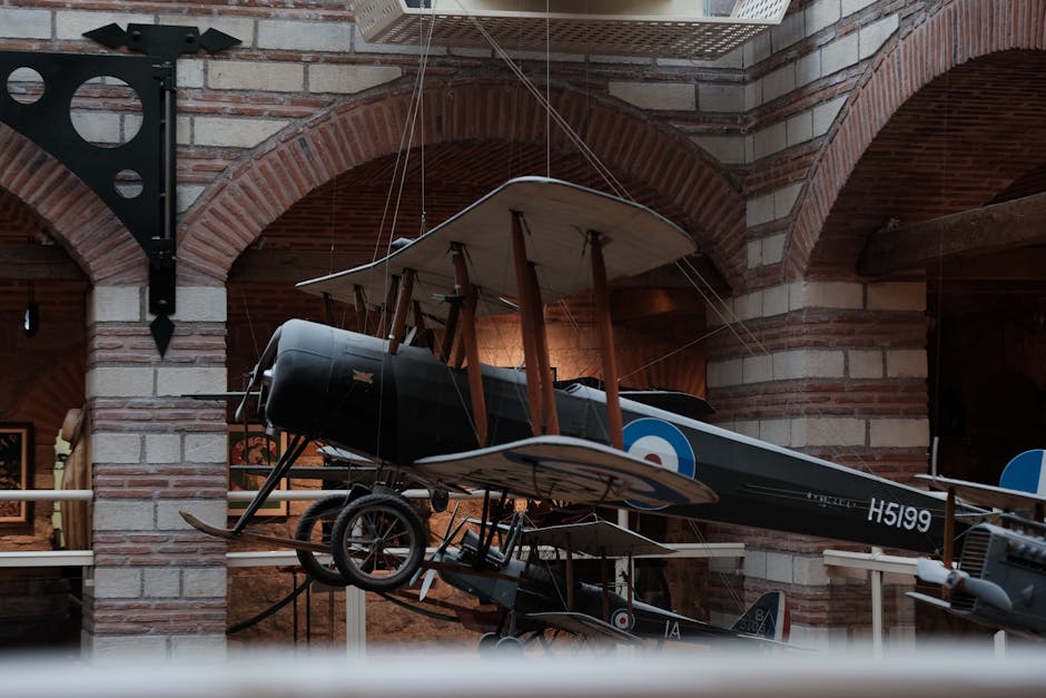 A vintage black and brown biplane aircraft with wooden propellers and fabric-covered wings is displayed indoors, suspended from the ceiling by thin wires. The aircraft features roundels on its fuselage, typical of military or historic aircraft, and registration markings visible beneath the wing. The interior setting includes brick arches and walls, with lighting highlighting the plane's structure and details. Surrounding elements include a glass barrier at waist height and informational plaques, suggesting the location is a museum, likely related to aviation or history. The aircraft is positioned centrally within the space, with the aircraft's nose pointing slightly downward and the tail elevated, indicating an exhibit on display. As part of the move or packing process, Man With a Van Hendon may be involved in transporting or installing such artifacts during home relocations or moving logistics near the Hendon landmark, supporting the visibility of museum or private collection items being prepared or displayed.