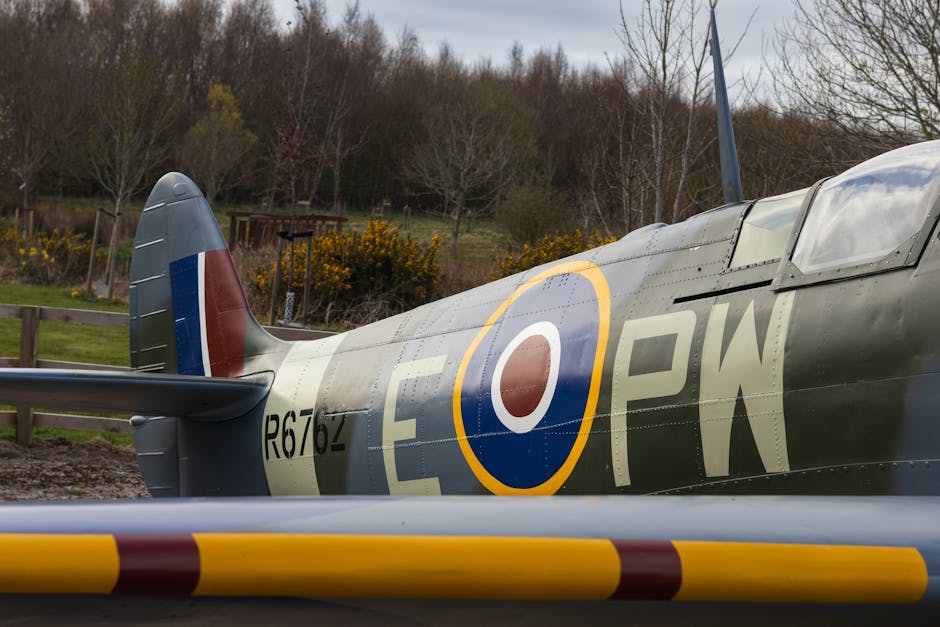 A vintage military aircraft with a silver-grey metallic fuselage is shown parked outdoors on a paved area. Visible features include the tail fin with blue, white, and red stripes, and a large roundel insignia with red, white, blue, and yellow colors on the side of the body. The aircraft's registration number R676ZP is painted in black near the tail section, along with additional markings. The cockpit canopy appears clear and is closed, with visible panels and rivets on the aircraft's surface. Surrounding the aircraft, there are yellow and brown striped safety barriers on the ground, and the setting includes a background of leafless trees, yellow flowering bushes, and overcast skies, indicating a rural or park environment. The scene is well lit with natural daylight, suggesting preparation or inspection for a possible event or display, relevant to the context of transporting or relocating historic aircraft as part of a museum or collection, similar to services offered by Man With a Van Hendon in the area near Hendon landmarks like the RAF Museum.