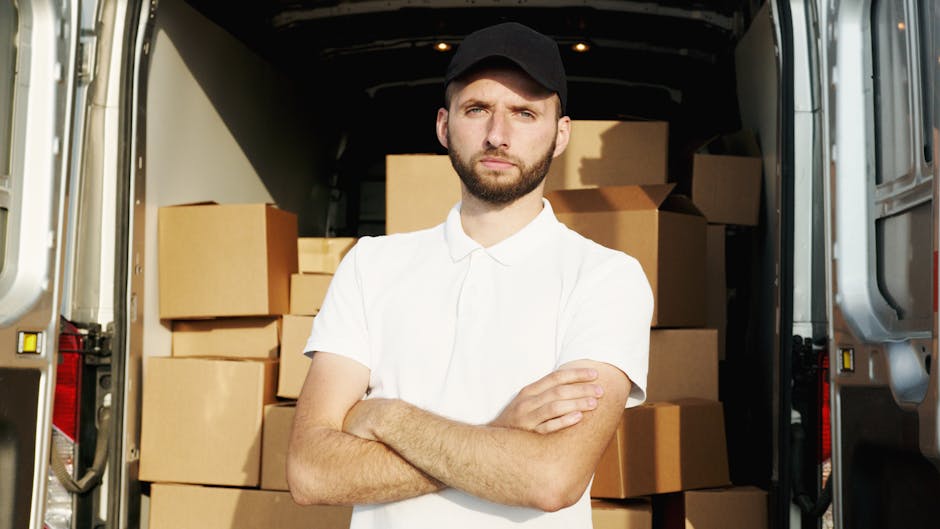 Two men wearing casual clothing and caps are engaged in a home relocation process, loading a large cardboard box into the back of a white van parked on a paved driveway. The van's rear doors are open,