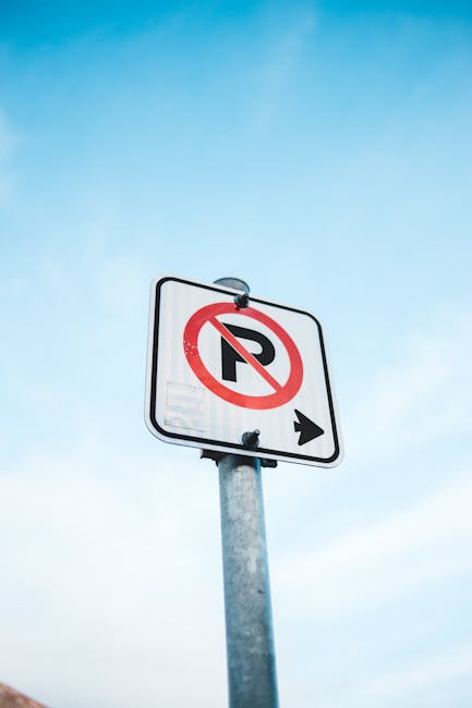 A vertical metal pole supporting a rectangular white traffic sign with a bold black ‘P’ crossed out by a red circle and slash, indicating no parking, with a black arrow pointing to the right at the bottom. The sign is mounted outdoors against a clear blue sky background. The image is related to parking regulations in Hendon and may be seen near residential or commercial areas used for house removals and furniture transport. Man With a Van Hendon ensures moving logistics adhere to local waste and skip rules, as discussed on their website, manwithavanhendon.co.uk.