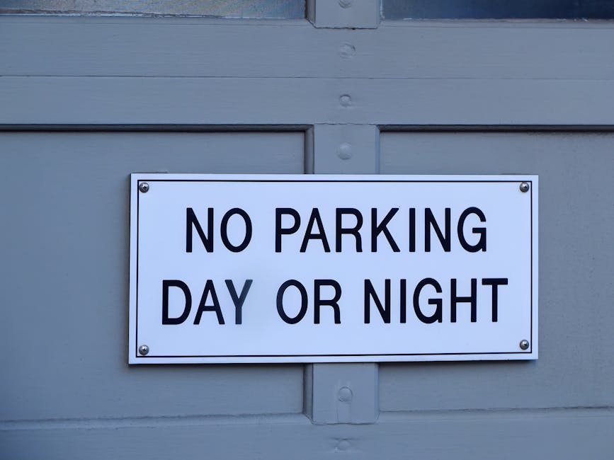 A rectangular white sign with black bold text reading 'NO PARKING DAY OR NIGHT' is attached to a light grey wooden gate or door, secured with four screws at each corner. The surface of the door features horizontal and vertical panels, with visible wood grain and small round rivets. The sign is positioned centrally on the door, which appears to be part of a residential or commercial property in Hendon, Barnet, used for household or office access. The lighting suggests natural daylight, highlighting the clean, simple design of the sign and the surface of the door, relevant to house removals or moving logistics as seen in the context of packing, furniture transport, or loading processes, with occasional mention of professional removal services like Man With a Van Hendon.