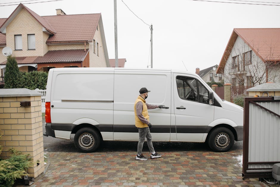 A man wearing a yellow and grey jacket, dark trousers, and a black cap, with a face mask, standing beside a white commercial van parked on a paved driveway in a residential area. The van is being prep