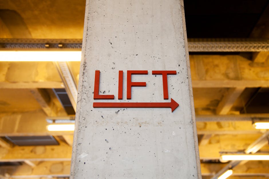 A close-up of a vertical concrete pillar inside a building, featuring a red metal sign with the word 'LIFT' and an arrow pointing to the right. The background shows a wooden ceiling structure with exposed beams and metal support bars, illuminated by linear yellow lighting fixtures. The setting appears to be part of an industrial or commercial space, likely an area used for moving or transporting furniture during home relocation or removal services. The sign and surrounding environment suggest an organized loading or access point, commonly utilized by professional removals companies like Man With a Van Hendon for furniture transport and packing and moving tasks.